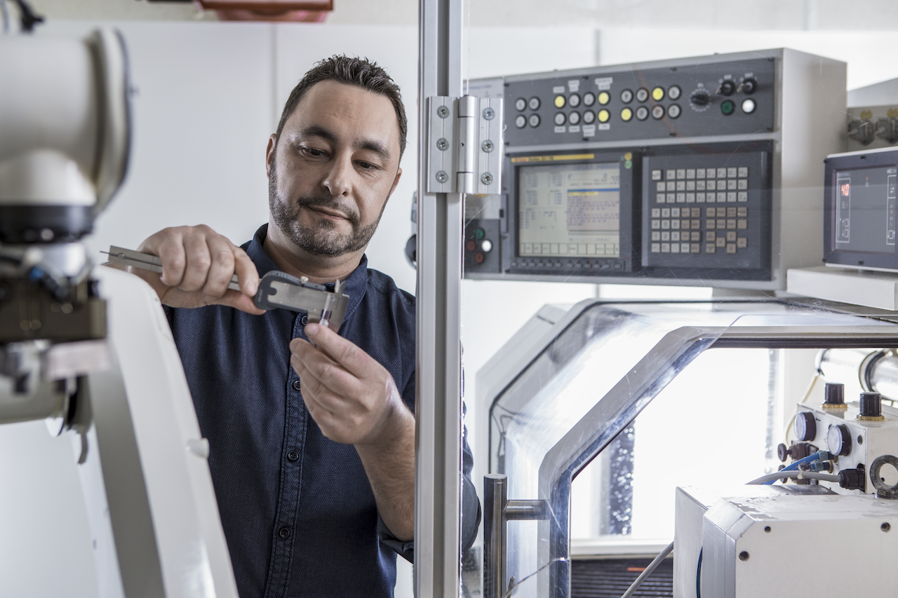 A technician uses a caliper to measure a component inside a tech lab, with advanced machinery and control panels in the background.